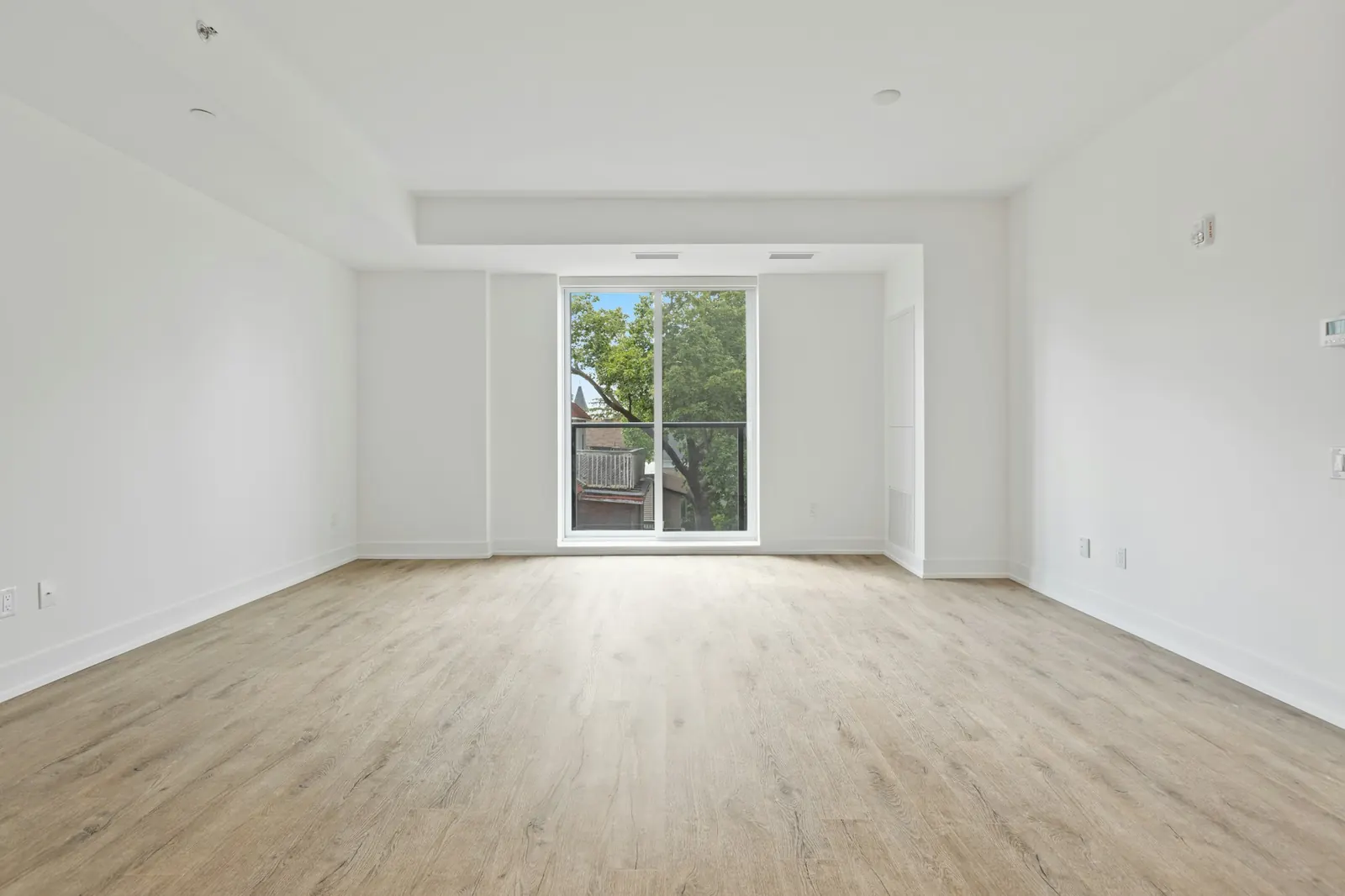 A vacant living room with pale oak floors and a tall window